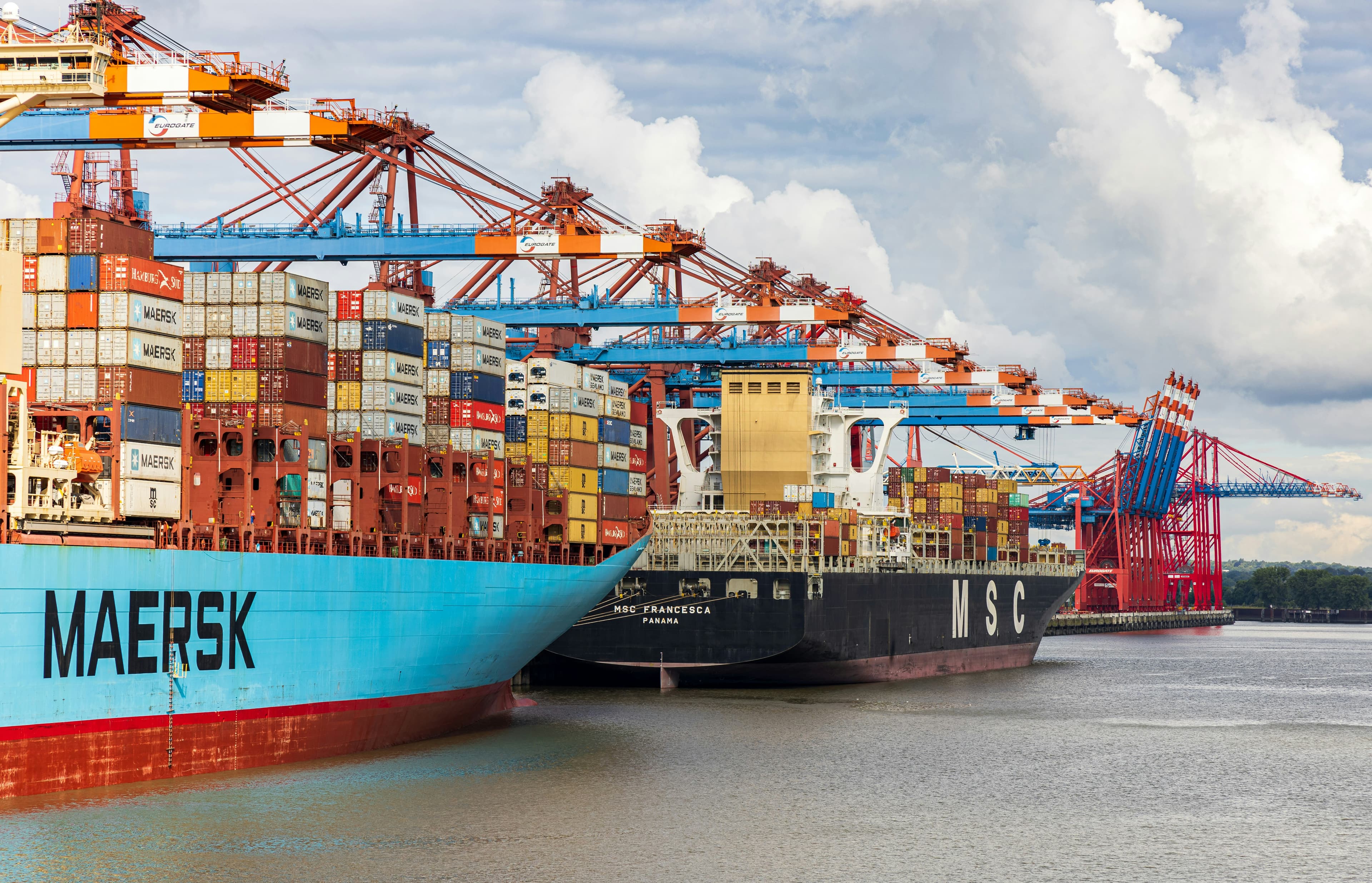Maersk and MSC container ships docked at a busy port with towering industrial gantry cranes.